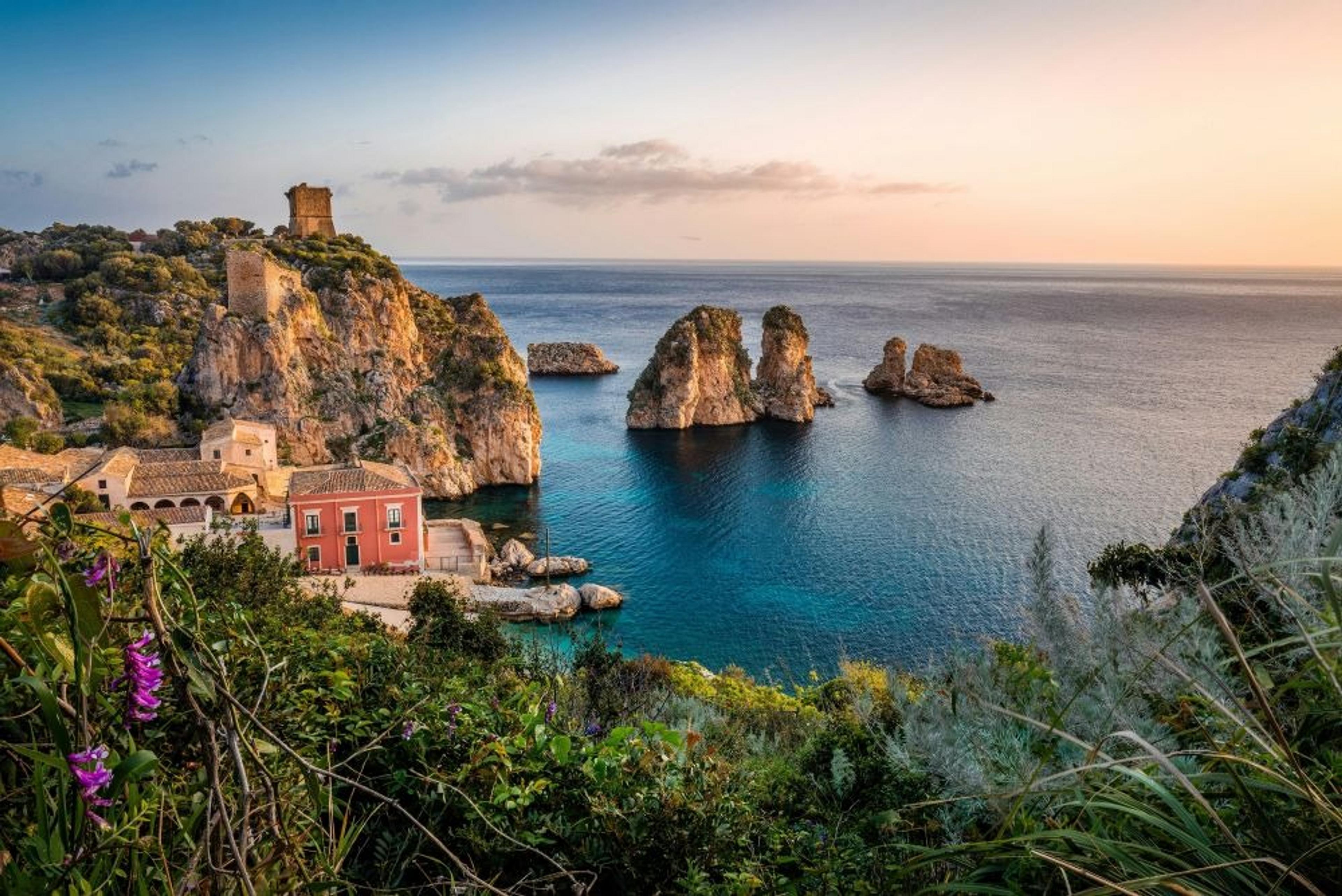 Cliffs and rock formations against a turquoise sea with a pink building and greenery in Sicily