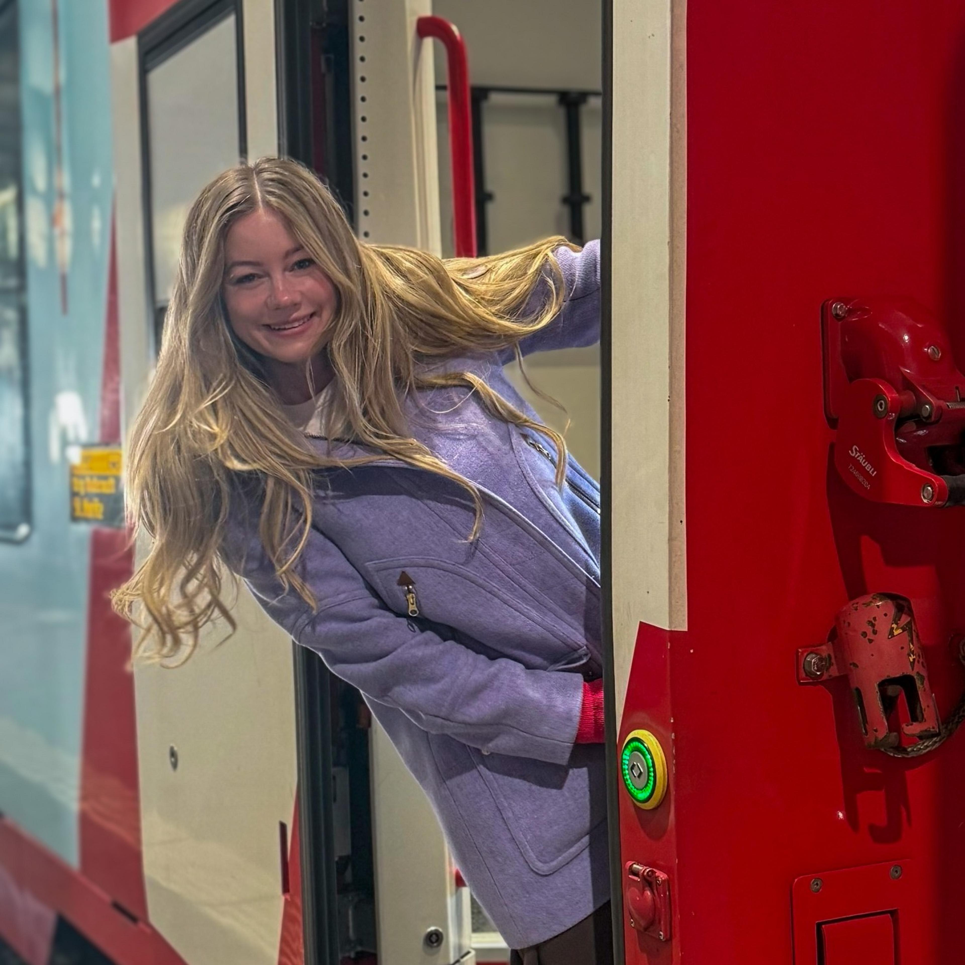 Kristi in a light purple coat leans out of a train door.